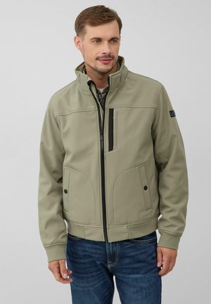 Veste softshell khaki avec un col haut, fermeture éclair à l'avant, accents noirs et deux poches latérales, associée à un jean bleu. Texture lisse.