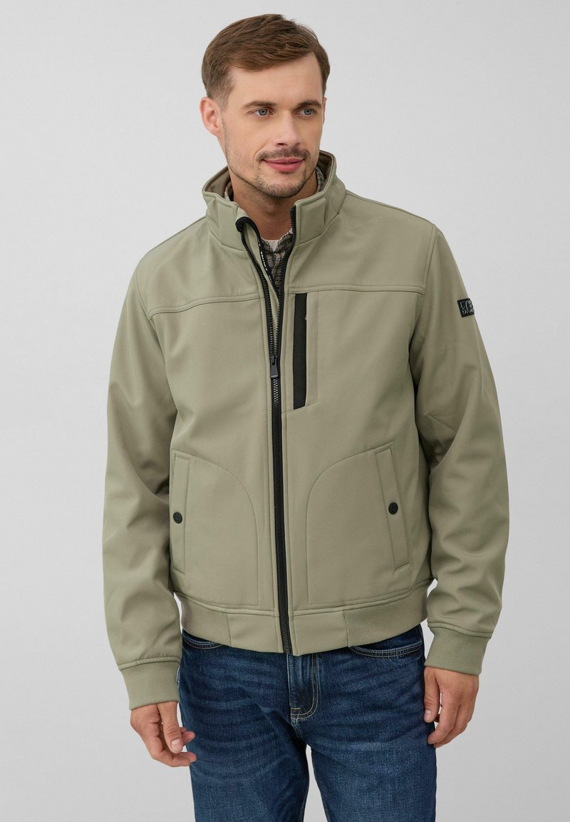 Veste softshell khaki avec un col haut, fermeture éclair à l'avant, accents noirs et deux poches latérales, associée à un jean bleu. Texture lisse.