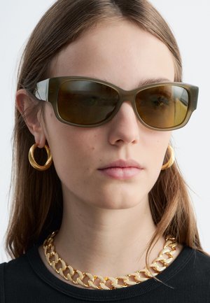 Jeune femme portant de grandes lunettes de soleil marron, de grosses boucles d'oreilles dorées en forme d'anneau, un collier chaîne doré épais et un haut noir avec des cheveux raides.