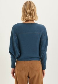 Blauwe gebreide trui met een brede ronde hals, verlaagde schouders en textuurpatronen. G gedragen met bruine hoge taillebroek.
