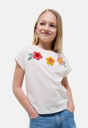Jeune fille aux cheveux blonds portant un t-shirt blanc avec des motifs de fleurs rouges, jaunes et roses, et un jean bleu, les mains dans les poches, souriante.