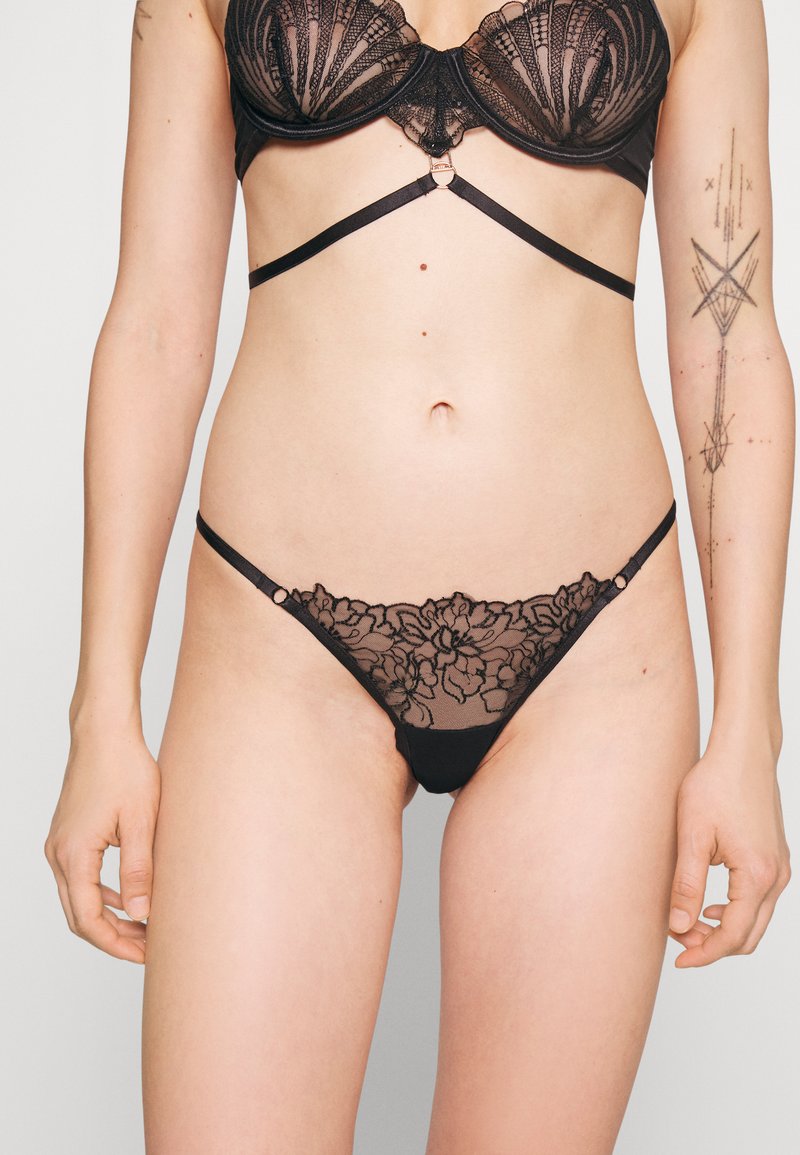 Conjunto de lingerie em renda preta com um sutiã balconette decorado com bordados florais e um fio dental transparente a condizer com correias decorativas.