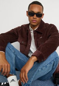 Pruunid viigipüksid, millel on kaks rinna taskut, kantud valge särgi peal. Heleda sinised teksad ja mustad prillid täiendavad välimust. Valged sokid on nähtavad.