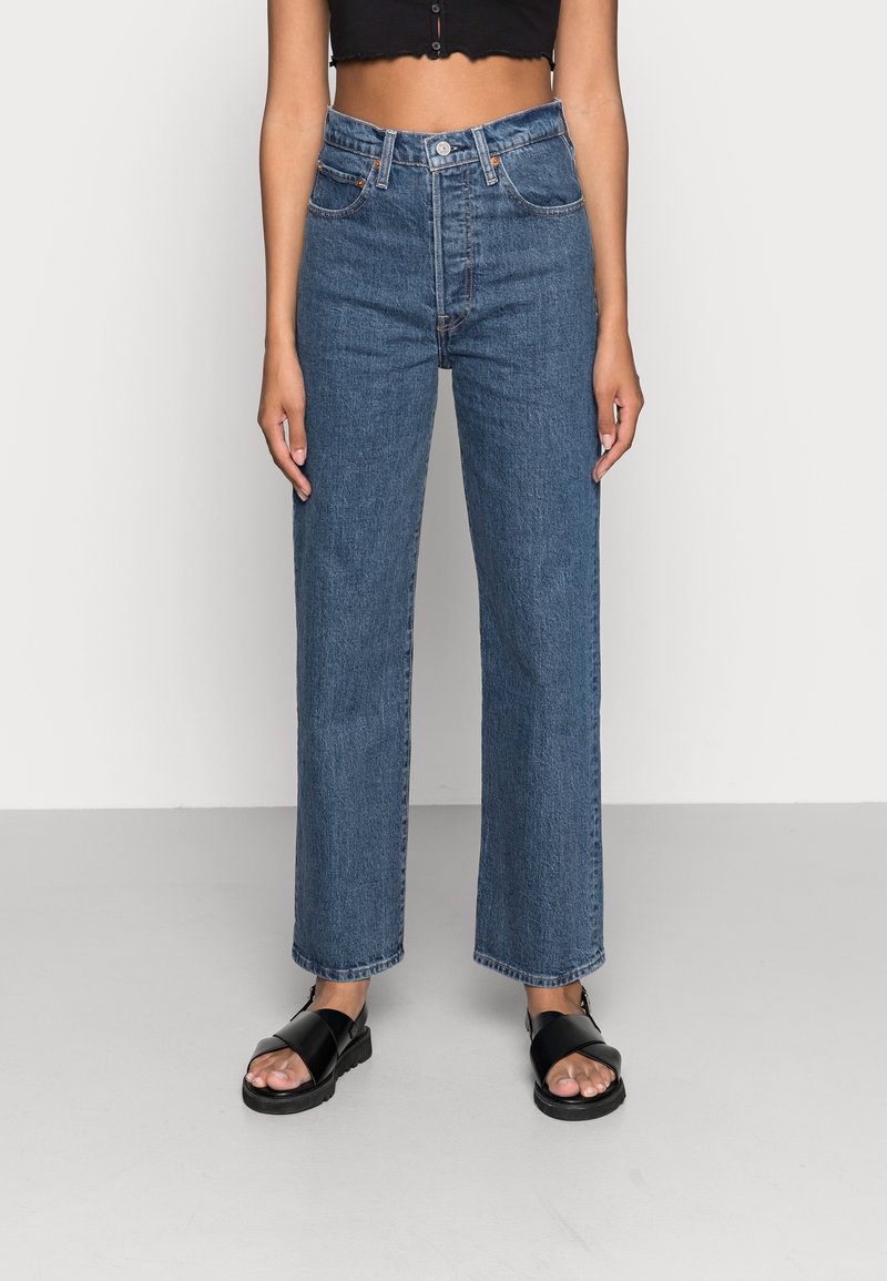 Personne portant un jean bleu taille haute à jambes droites, un crop top noir et des sandales noires à bout ouvert, debout devant un fond uni.