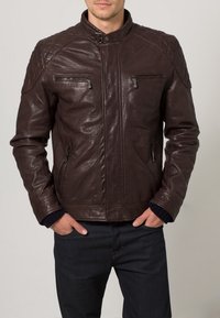 Hnědá kožená bunda s vysokým límcem, prošitými ramenními detaily, zipovanými kapsami na hrudi a bocích a žebrovanými manžetami. Hladká textura.