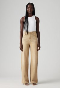 Μπεζ τζιν παντελόνι με φαρδιά γραμμή και ψηλή μέση, με ραμμένα στοιχεία και διακριτική υφή, σε συνδυασμό με ένα εφαρμοστό λευκό αμάνικο τοπ.
