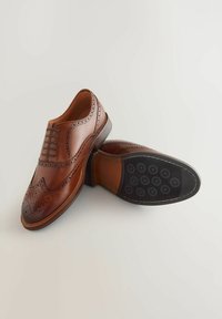 Pantofi din piele maro, cu design tip wingtip, detalii perforate și o finisare netedă. Talpa din cauciuc negru cu modele circulare.