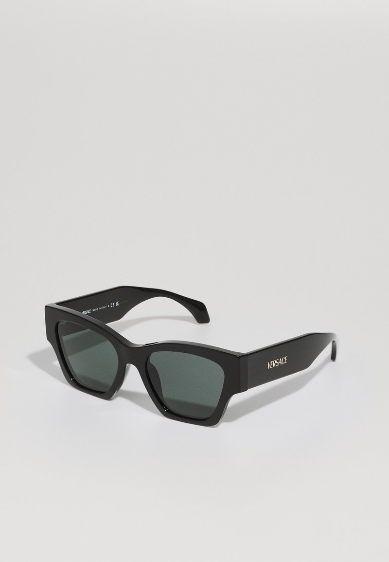Μαύρα γυαλιά ηλίου με γεωμετρικό σχήμα γατίσιας ματιάς, ματ φινίρισμα, σκουρόχρωμοι φακοί και χρυσή σήμανση "Versace" στο σκουρό.