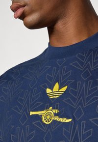 Camiseta de algodón de color azul marino con un patrón geométrico texturizado, logo de Adidas bordado en amarillo y diseño de cañón en el pecho.