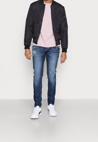 Černá bomber bunda, světle růžové tričko, roztrhané modré džíny a bílé tenisky. Detaily zahrnují roztržení na džínech a kapsu na zip na bundě.