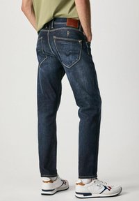 Vaqueros de mezclilla azul oscuro con un diseño de pierna recta, que cuentan con bolsillos traseros y un parche de cuero marrón en la cintura. Zapatillas blancas completan el look.