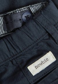 Mörkblå denimtyg med ett randigt invändigt midjeband, utrustad med en mörk plastknapp och en vit etikett som säger "newbie."