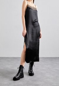 Robe nuisette en satin noir avec des bordures en dentelle, dotée de fines bretelles spaghetti et de fentes sur les côtés. Assortie à des bottes de combat noires à lacets.