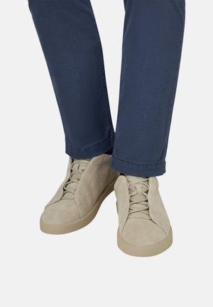 Baskets en daim beige avec lacets portées avec un pantalon bleu foncé sur un fond clair uni.