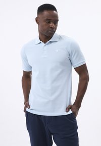 Helles blaues Poloshirt aus strukturiertem Stoff, mit Kragen, kurzen Ärmeln und einem dezenten Logo auf der linken Brust.