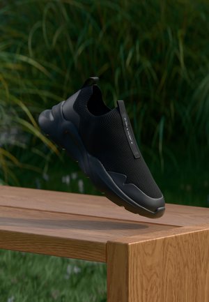 Sneaker nera senza lacci con tomaia in mesh testurizzato, punta tonda, suola spessa e imbottita, e un pratico passante. Ambientata su uno sfondo verde naturale.