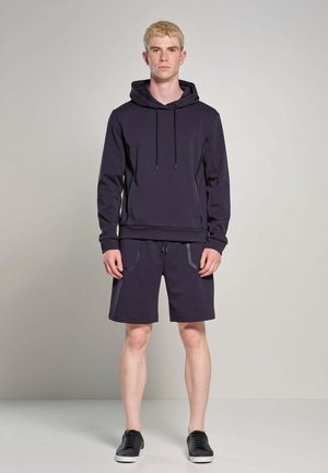 Jonge man met blond haar, gekleed in een marineblauwe capuchontrui en bijpassende korte broek, staand tegen een eenvoudige lichte achtergrond.
