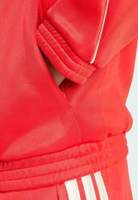 Red track jacket features ribbed cuffs and waistband, smooth texture, with white stripes accenting the sides. Includes a zippered pocket.