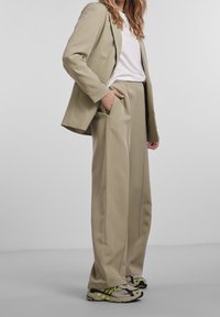 Femme portant un pantalon large beige, un blazer assorti, un t-shirt blanc, et des baskets grises avec des accents vert néon, debout devant un fond uni.