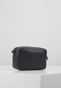 Černá kožená taštička na toaletní potřeby s obdélníkovým tvarem, zapínáním na zip, bočním úchopem a hladkou texturou. Minimalistický design bez viditelných vzorů.