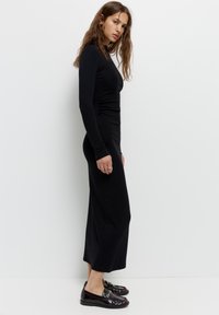 Rochie neagră cu mâneci lungi, cu un corset ajustat, pliseuri laterale și o siluetă fluidă. Asortată cu mocasini negri lucioși, cu o cataramă din argint.