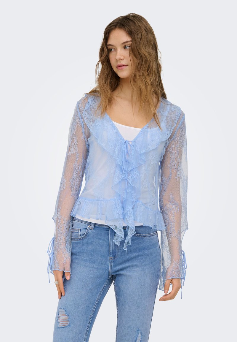 Jeune femme porte un chemisier en dentelle bleu clair transparent avec des volants sur un top blanc, associé à un jean bleu déchiré, debout devant un fond uni.