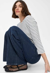 Modell trägt ein weißes, langärmliges gestreiftes Oberteil und blaue weite Jeans. Schwarze Mesh-Ballettschuhe und ein silbernes Armband sind sichtbar.