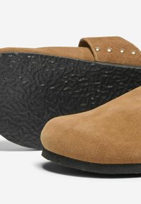Scarpe slip-on in suede marrone con suola in gomma nera testurizzata, dotate di una fascia decorata con borchie metalliche. Design a punta arrotondata.