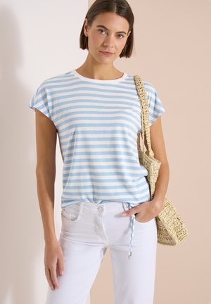 Femme portant une chemise à manches courtes rayée bleu clair et blanc, un pantalon blanc, tenant un sac en paille tressée sur son épaule.