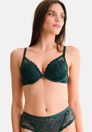 Soutien-gorge en dentelle vert foncé avec des bords festonnés et des motifs floraux, équipé de bretelles ajustables et d'une fermeture à agrafe. Modèle portant des culottes assorties.
