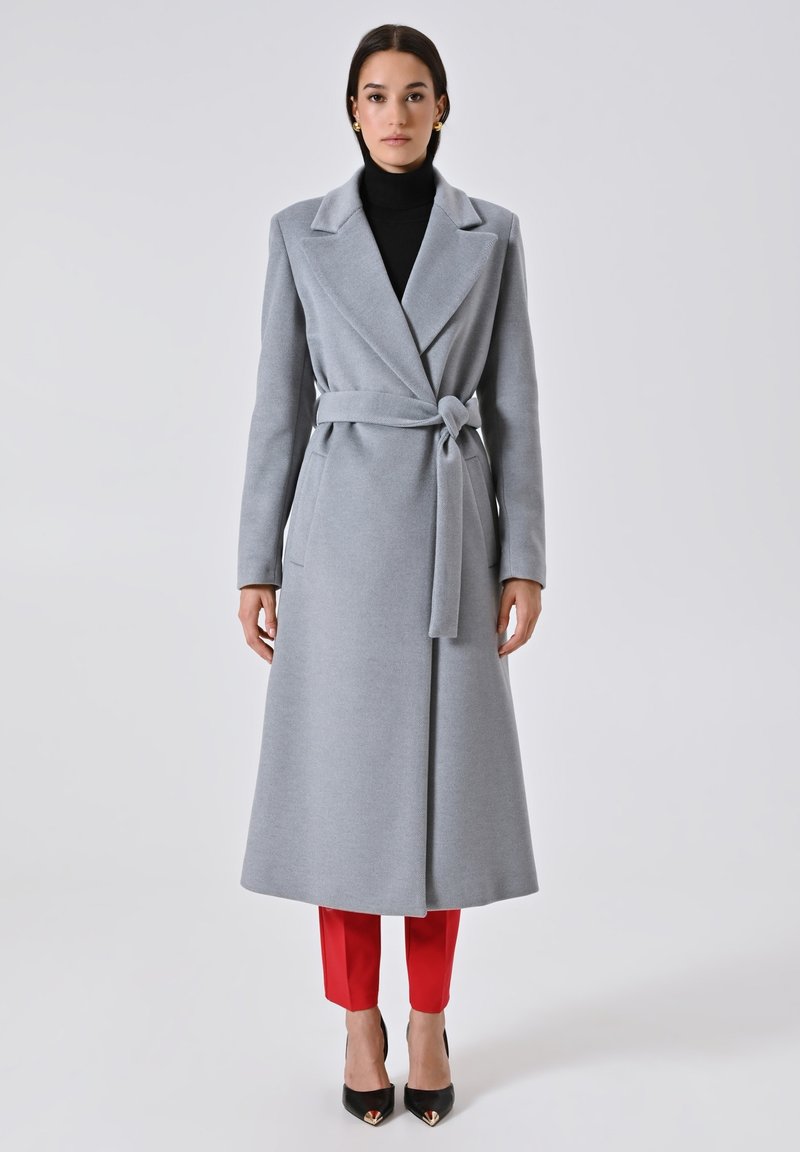 Grauer Wollmantel mit Wickeldesign, breiten Revers und Bindegürtel, kombiniert mit roten, cropped Pants und schwarzen spitzen Pumps.
