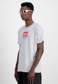 Siva kratka majica s kratkimi rokavi z rdečim in �črnim logotipom "ALPHA INDUSTRIES", zasnova s krožnim izrezom in majhna črna oznaka na rokavu.