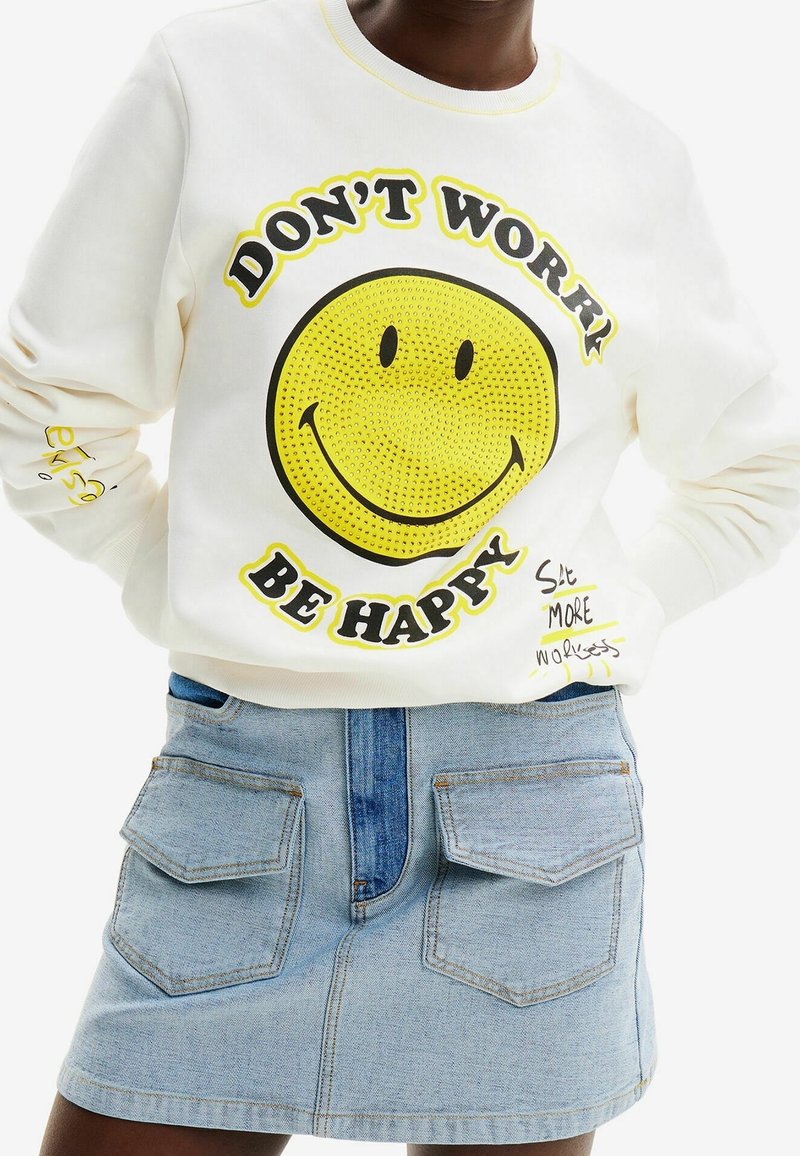 Sweat-shirt avec un grand visage souriant jaune et le texte « DON'T WORRY BE HAPPY », assorti à une jupe en jean bleu clair avec des poches avant.