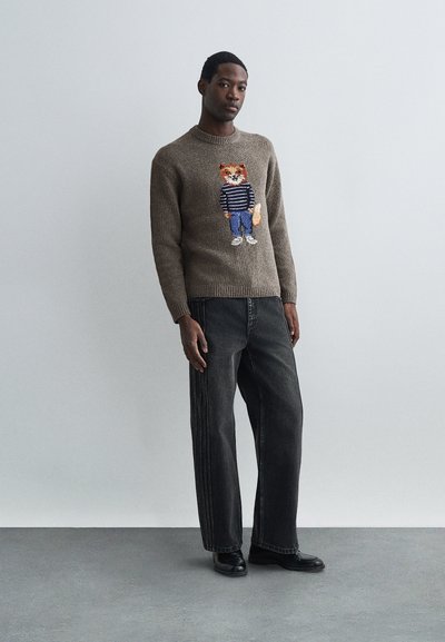 Vīrietis, kas ģērbies brūnā džemperī ar karikatūras lapsu, svītrainā krekla un džinsos, stāv uz pelēka grīdas pretī vienkāršai pelēkai sienai.