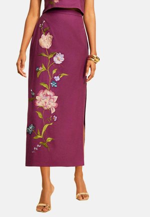 Frau trägt einen lila Midi-Rock mit Blumenstickerei auf der Vorderseite, kombiniert mit goldenen offenen Schuhen und klobigen goldenen Armbändern an einem Handgelenk.