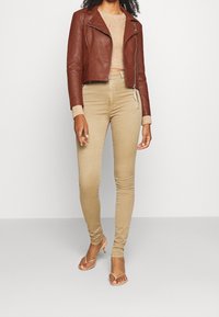 Kobieta w brązowej skórzanej kurtce, beżowym krótkim swetrze dzianinowym, beżowych dopasowanych spodniach i beżowych sandałach na obcasie z odkrytymi palcami.