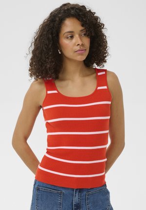 Kohutüdruk, kes kannab punast triibulist varrukateta topi ja siniseid teksaseid, seisab ühevärvilise tausta ees.