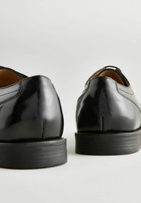 Schwarze Lederschuhe mit glänzender Oberfläche, runder Zehenpartie und einer niedrigen schwarzen Gummisohle. Nähte sind entlang der Teilungsnähte sichtbar.