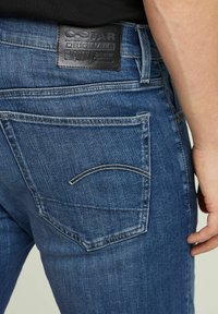 Jeans din denim albastru cu o suprafață texturată. Prezintă un etichete din piele pe talie și un buzunar pe spate cu un detaliu de cusătură curbată distinct.