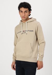 Ανοιχτό μπεζ φούτερ από μαλακό ύφασμα, με μπροστινή τσέπη, κουκούλα με κορδόνι και ναυτικό λογότυπο με κόκκινες και λευκές λεπτομέρειες.