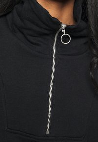 Sweat-shirt noir à fermeture éclair avec un col montant. Il présente une fermeture éclair argentée avec un anneau circulaire comme tirette. Texture côtelée sur le col et les poignets.