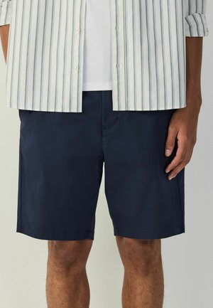 Mand iført navyblå shorts og en hvid skjorte med blå lodrette striber, står med hånden afslappet ned langs siden.