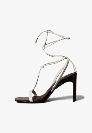 Weiße Riemchensandalette mit hohem Absatz und quadratischer Spitze, glatten Lederriemen und einer braunen, strukturierten Innensohle. Bänder wickeln sich um den Knöchel und enden in metallischen Spitzen.