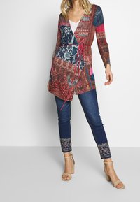 Mönstrad cardigan i rika röda, blå och rosa nyanser, med trekvartsärmar och midjeband. Matchad med mörkblå jeans med broderi.