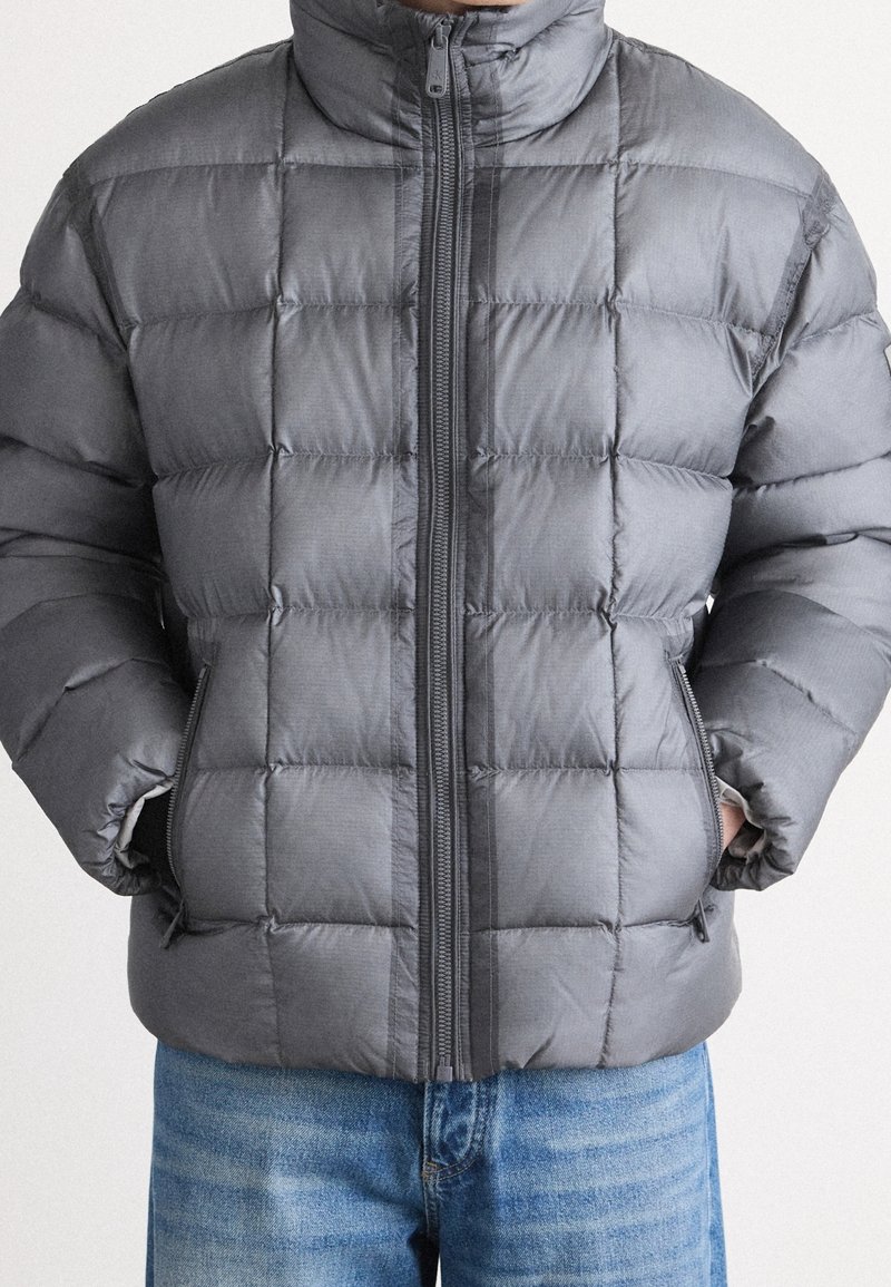 Personne portant une doudoune matelassée grise zippée, les mains dans les poches avant, et un jean bleu visible à partir du milieu des cuisses.
