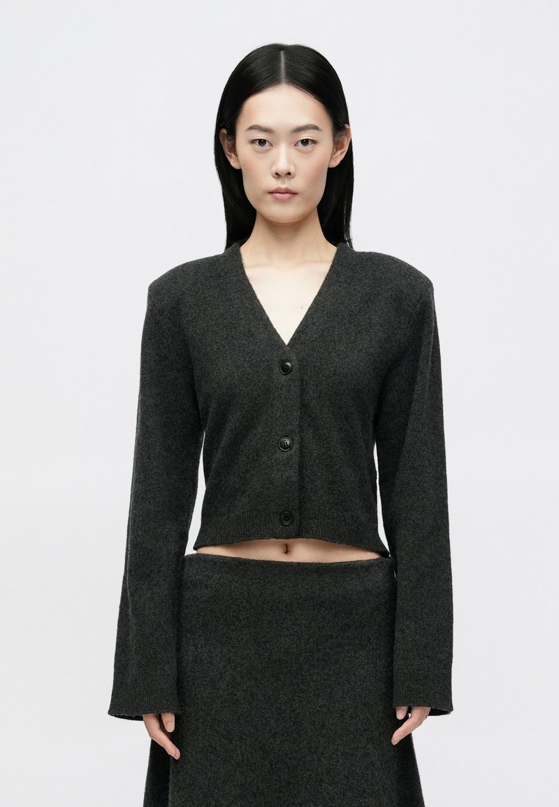 Γυναίκα με ίσια μαύρα μαλλιά φοράει σκούρο γκρι πουλόβερ με κουμπιά και αντίστοιχη φούστα, στέκεται μπροστά σε απλό ανοιχτό γκρι φόντο.