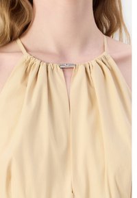Krémový halter top s prešívanou látkou na výstrihu a strieborným uzáverom vpredu. Hladká textúra, ľahký materiál.