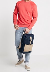 Granatowy i beżowy plecak z regulowanymi ramiączkami, przednią kieszenią i zapięciem na klamrę. Nosi się go z koralowym swetrem i jasnoniebieskimi dżinsami.