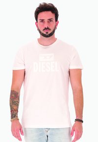 Giovane uomo con barba e orecchini che indossa una maglietta bianca Diesel, jeans chiari, braccio sinistro tatuato e braccialetti a entrambe i polsi, in piedi contro uno sfondo bianco.