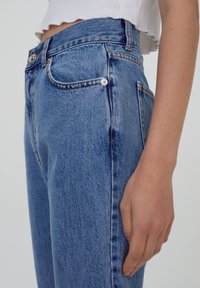 Jean en denim bleu clair à taille haute, avec un design à cinq poches, un léger délavage et des coutures classiques. Comprend une fermeture à bouton à l'avant et des passants de ceinture.